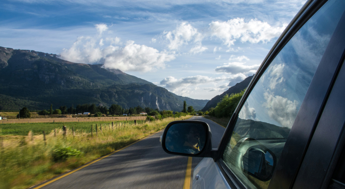Car driving along a country road through a green landscape with mountains in the background, seen from the side mirror.