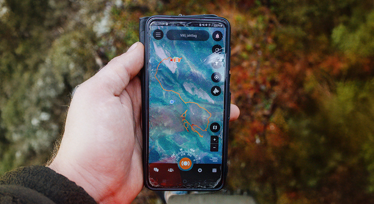 En jägare håller sin mobil ute i skogen. På mobilskärmen syns jaktappen MiniFinder Hunter med karta som visar hundens position under jakt med hjälp av GPS-spårning.