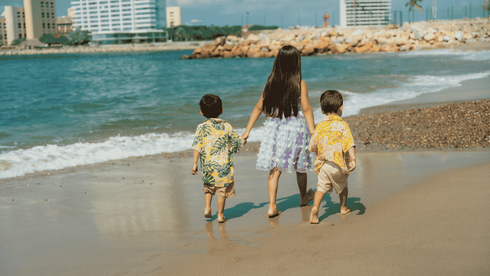 Three siblings walking hand in hand along a beach abroad while wearing a GPS watch for children.